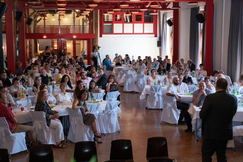 Ein weitläufiger Raum, in dem zahlreiche Personen an Tischen Platz genommen haben.