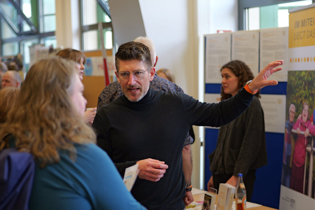 Ein Mann spricht mit Personen an einem Tisch, während ein Poster im Hintergrund sichtbar ist.