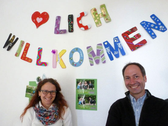 Zwei Menschen posieren vor einer Wand, auf der "lich willkommen" in großen Buchstaben steht.