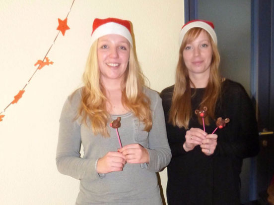 Zwei Frauen mit Weihnachtsmützen halten bunte Lutschbonbons in den Händen und lächeln fröhlich.