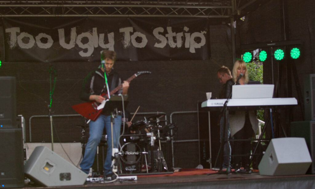 Live-Musik: Band "Too Ugly To Strip" auf der Bühne mit Gitarrist, Keyboarder und Sängerin bei einem Konzert.