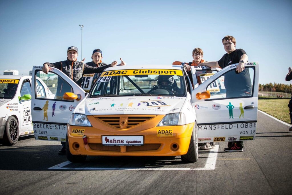 Motorsport-Team mit Rennwagen Dacia Logan beim ADAC Clubsport Rennen. Fahrer und Teammitglieder vor dem Auto auf der Rennstrecke.