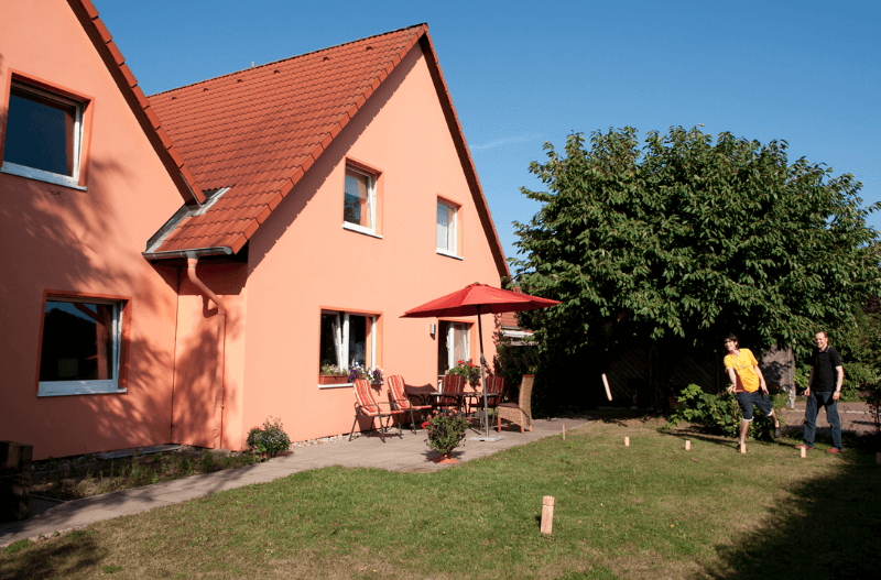 Haus mit Garten, Terrasse und spielenden Personen. Kubb-Spiel im Freien, Sommeraktivität. Familienspiel im Garten.