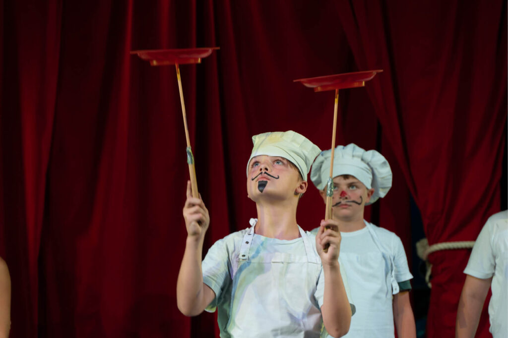 Kinderzirkus: Junge Artisten jonglieren mit Tellern auf Stäben vor rotem Vorhang. Zirkusaufführung mit Clowns und Akrobatik.