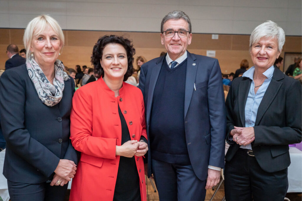 Politiker-Gruppe bei Veranstaltung: Vier Personen, zwei Frauen in Blazer, ein Mann im Anzug, Konferenz im Hintergrund.