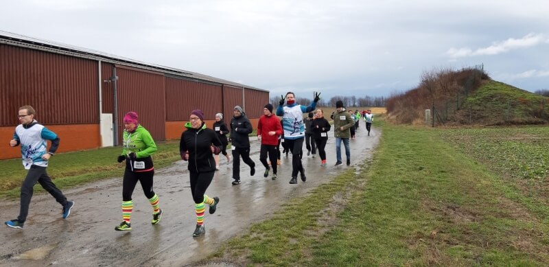 Laufveranstaltung: Gruppe von Läufern auf schlammiger Strecke, Gebäude und Hügel im Hintergrund. Outdoor-Sport bei jedem Wetter.