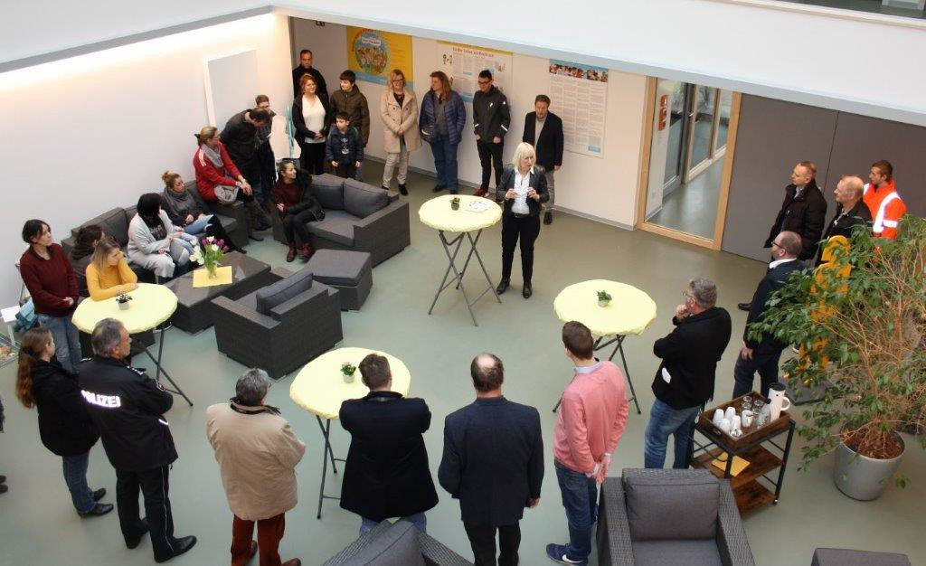 Eine Gruppe von Menschen steht und sitzt in einem modernen Innenraum, versammelt um kleine runde Tische und hört einer Frau zu, die in der Mitte spricht. Einige tragen Mäntel, und auf einem Beistelltisch stehen Erfrischungen.