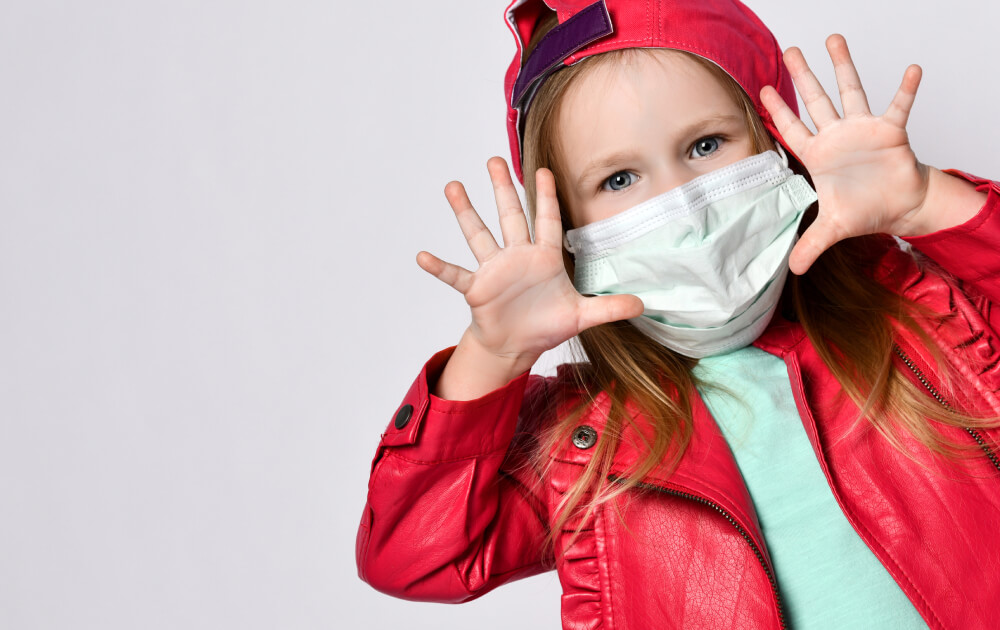 Ein kleines Kind, das eine rote Jacke, eine rote Mütze und eine medizinische Gesichtsmaske trägt, hält spielerisch beide Hände mit gespreizten Fingern vor einem schlichten hellen Hintergrund hoch.