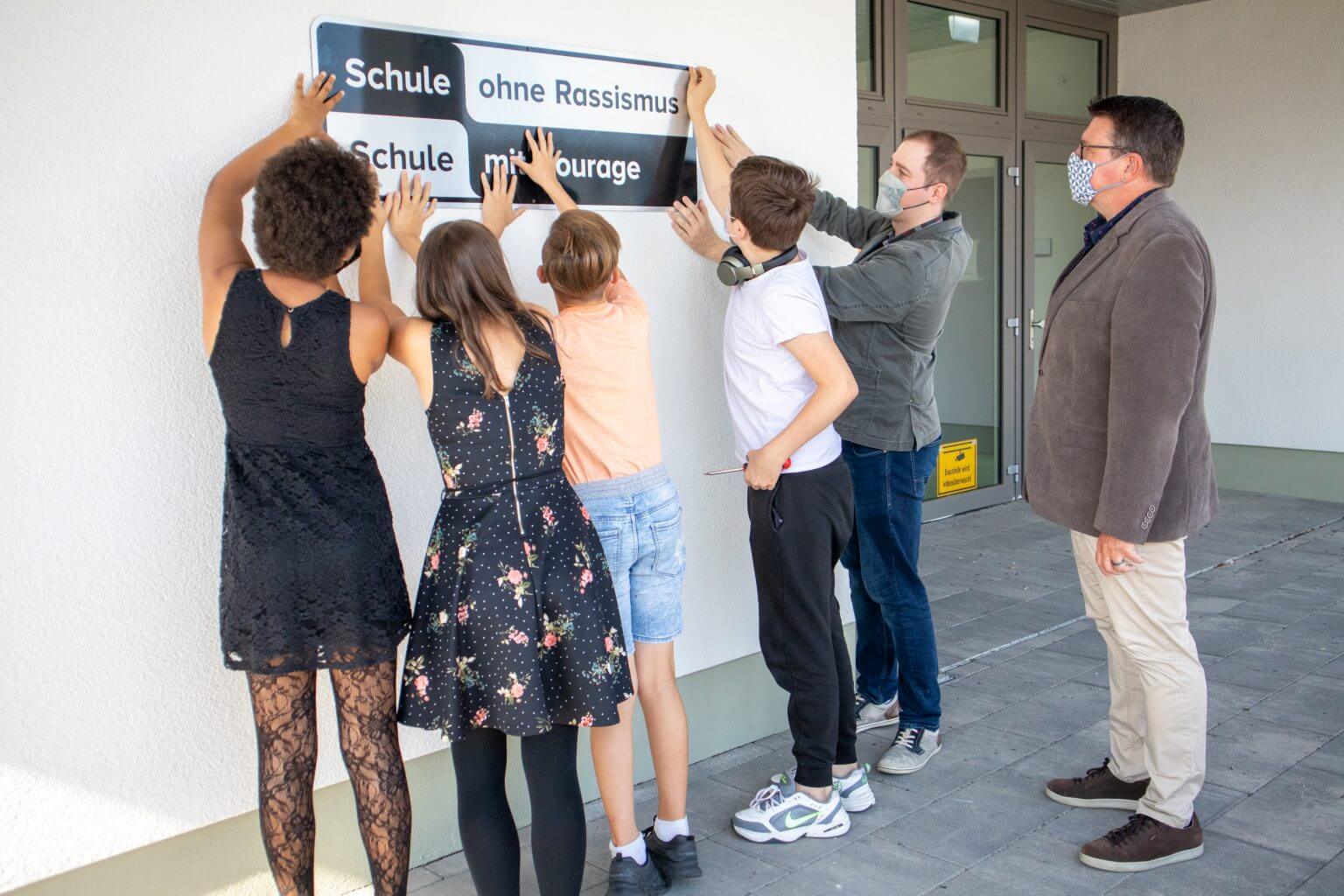 Fünf Personen, darunter Schüler und ein Erwachsener, befestigen ein Schild mit der Aufschrift "Schule ohne Rassismus, Schule mit Courage" an einer Wand. Der Erwachsene und ein Schüler tragen Gesichtsmasken. Alle Gesichter sind abgewandt oder zur Seite gedreht.