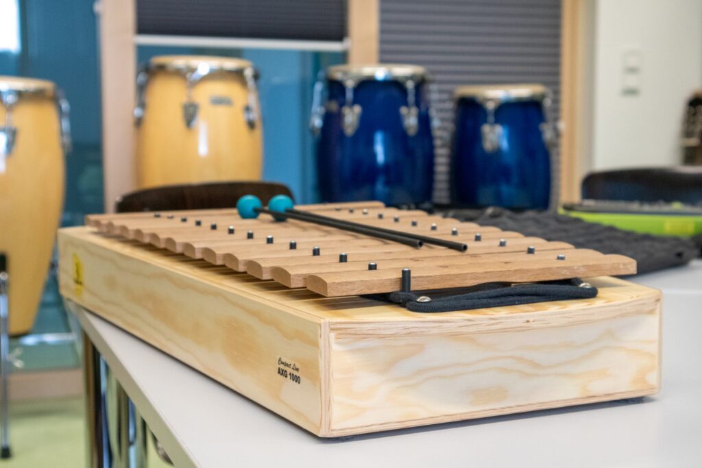 Ein hölzernes Xylophon mit zwei Schlägeln liegt auf einem Tisch in einem Musikzimmer; im Hintergrund sind blaue und gelbe Conga-Trommeln zu sehen.