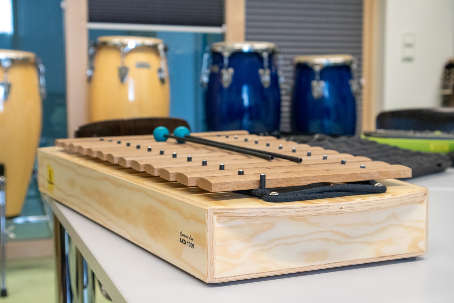 Ein hölzernes Xylophon mit zwei Schlägeln liegt auf einem Tisch in einem Musikzimmer; im Hintergrund sind blaue und gelbe Conga-Trommeln zu sehen.