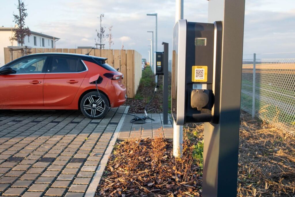 Ein rotes Elektroauto ist auf einem gepflasterten Parkplatz neben einem Holzzaun geparkt und an eine Ladestation angeschlossen, während im Hintergrund ein offenes Feld und ein Maschendrahtzaun zu sehen sind.