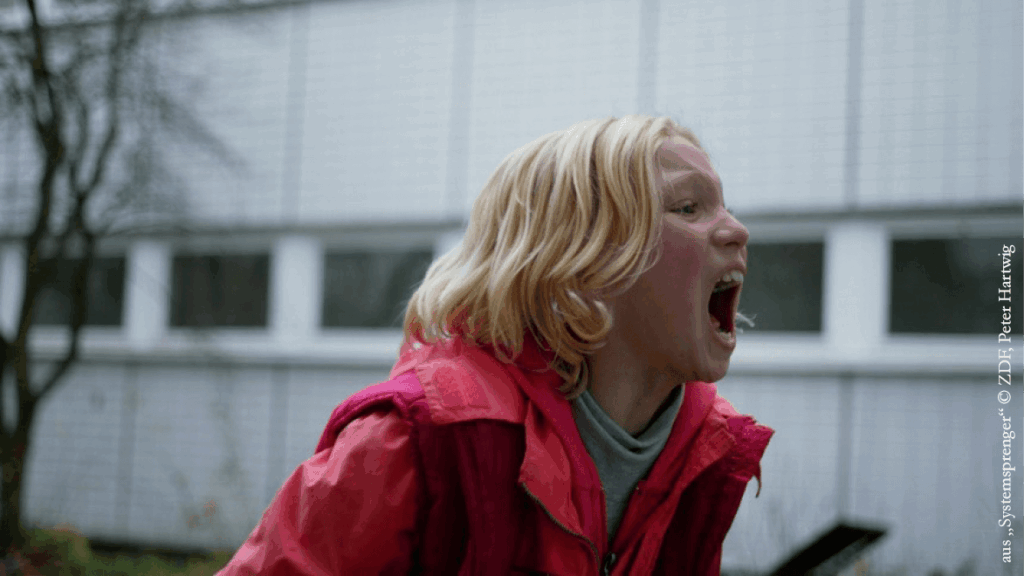 Eine junge Person mit blonden Haaren und einer hellrosa Jacke steht im Freien und schreit mit weit geöffnetem Mund. Im Hintergrund sind ein weiß gekacheltes Gebäude mit Fenstern und einige kahle Bäume zu sehen.