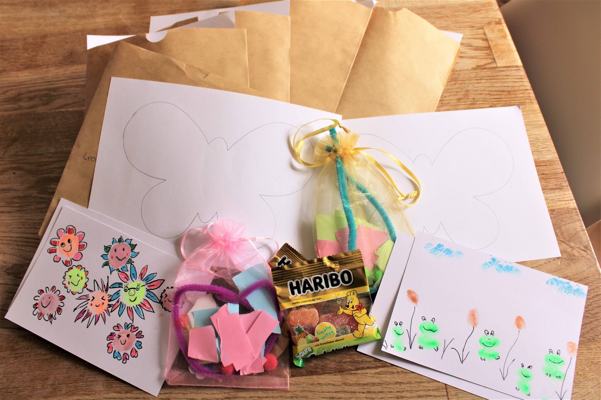 Auf einem Holztisch liegen Briefumschläge, Malblätter mit Schmetterlingen und Blumen, zwei verzierte Karten, Schaumstoffbasteleien in Netzbeuteln und eine kleine Packung Haribo-Gummibärchen.