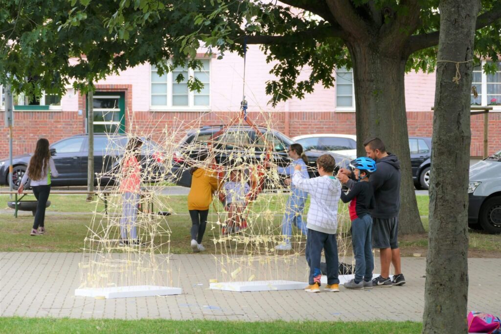 Kinder und Erwachsene versammeln sich im Freien um eine große Struktur aus miteinander verbundenen Stöcken oder Stäben, wahrscheinlich als Teil einer kreativen oder pädagogischen Aktivität, mit Bäumen und Gebäuden im Hintergrund.