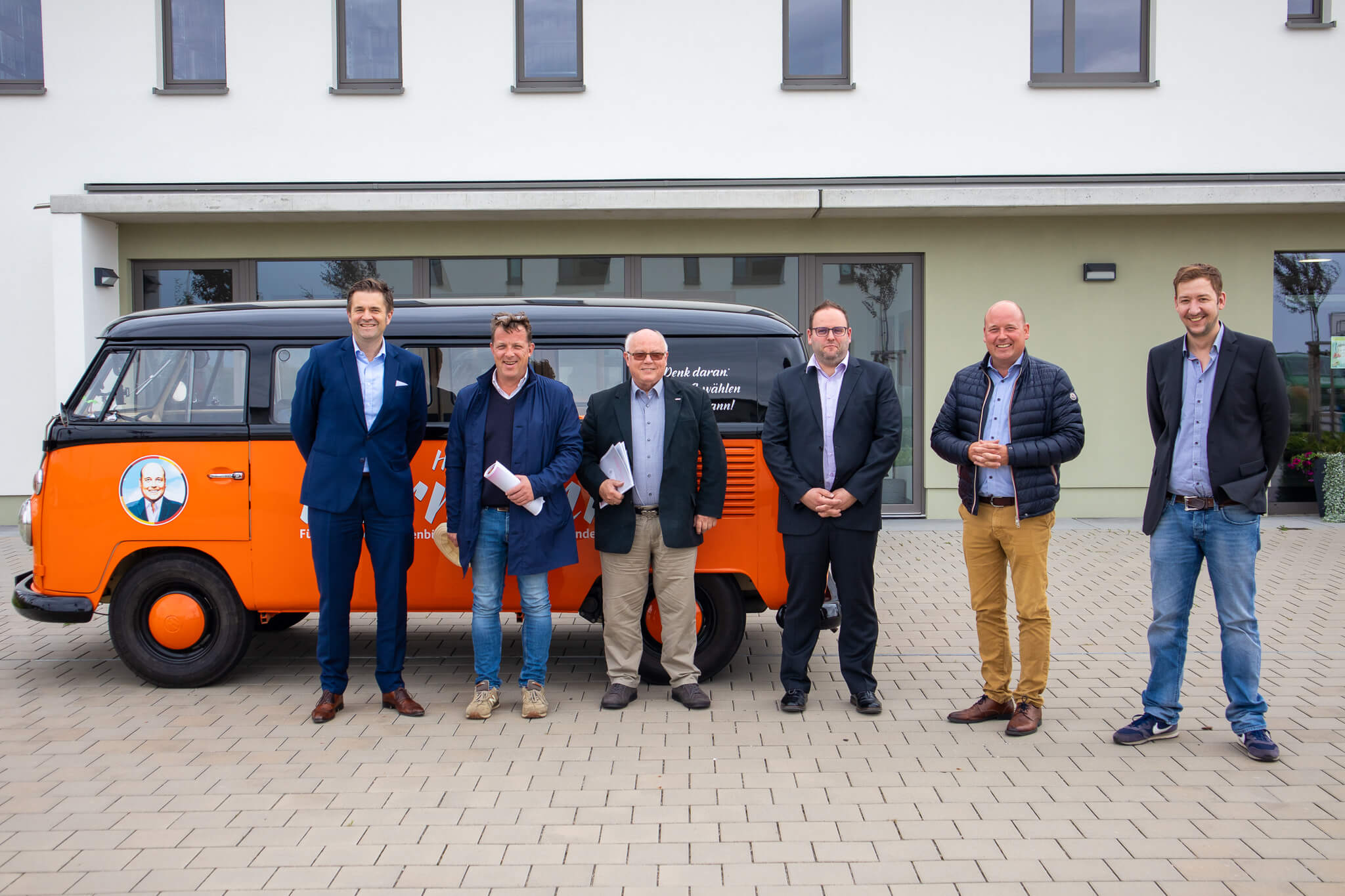 Sechs Männer stehen lächelnd vor einem orange-schwarzen Oldtimer-Van, der vor einem modernen weißen Gebäude mit großen Fenstern geparkt ist. Die Männer sind in eleganter Freizeit- und Geschäftskleidung gekleidet.
