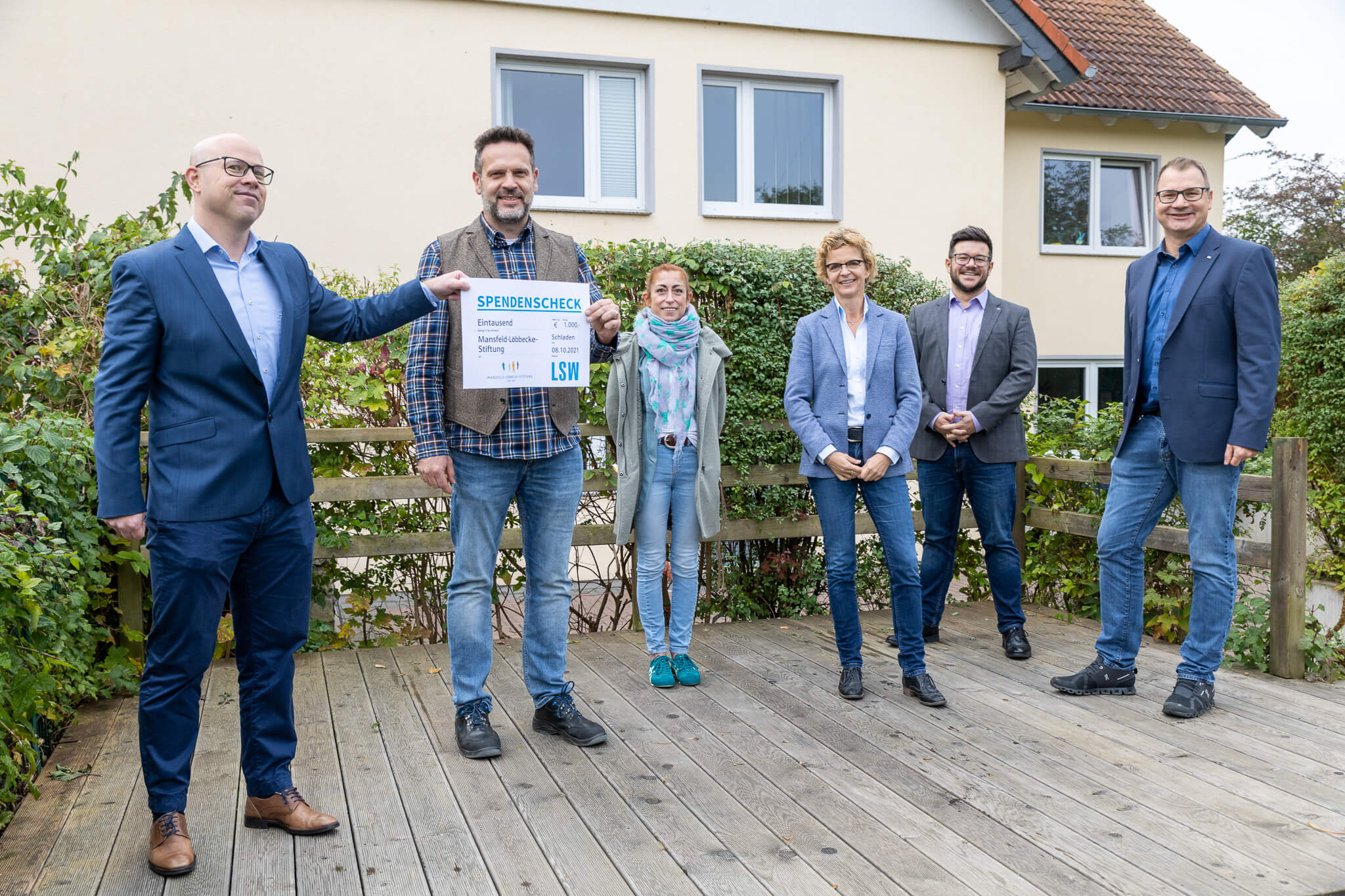 Sechs Personen stehen draußen auf einem Holzdeck und lächeln. Eine Person hält einen großen Spendenscheck in der Hand. Sie sind leger gekleidet und stehen vor einem Haus, das im Hintergrund grünt.