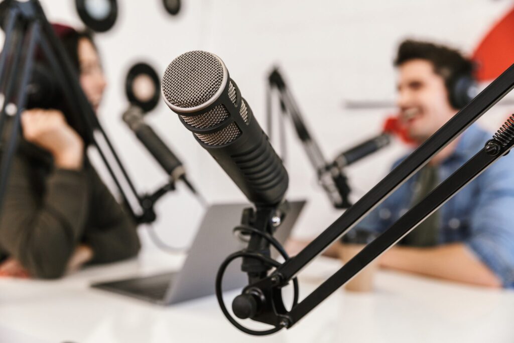 Nahaufnahme eines Mikrofons auf einem Ständer in einem Aufnahmestudio, mit zwei Personen mit Kopfhörern, die sich im unscharfen Hintergrund unterhalten, was auf einen Podcast oder ein Radiointerview hindeutet.