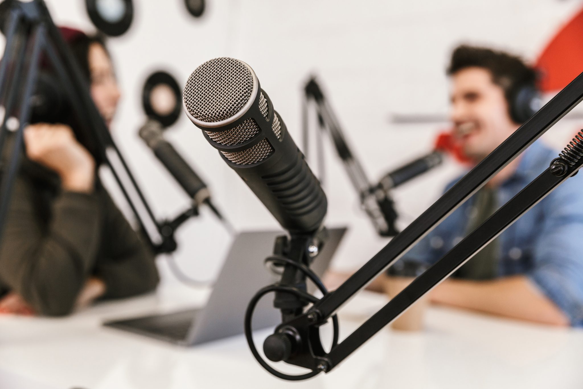 Nahaufnahme eines Mikrofons auf einem Ständer in einem Aufnahmestudio, mit zwei Personen mit Kopfhörern, die sich im unscharfen Hintergrund unterhalten, was auf einen Podcast oder ein Radiointerview hindeutet.