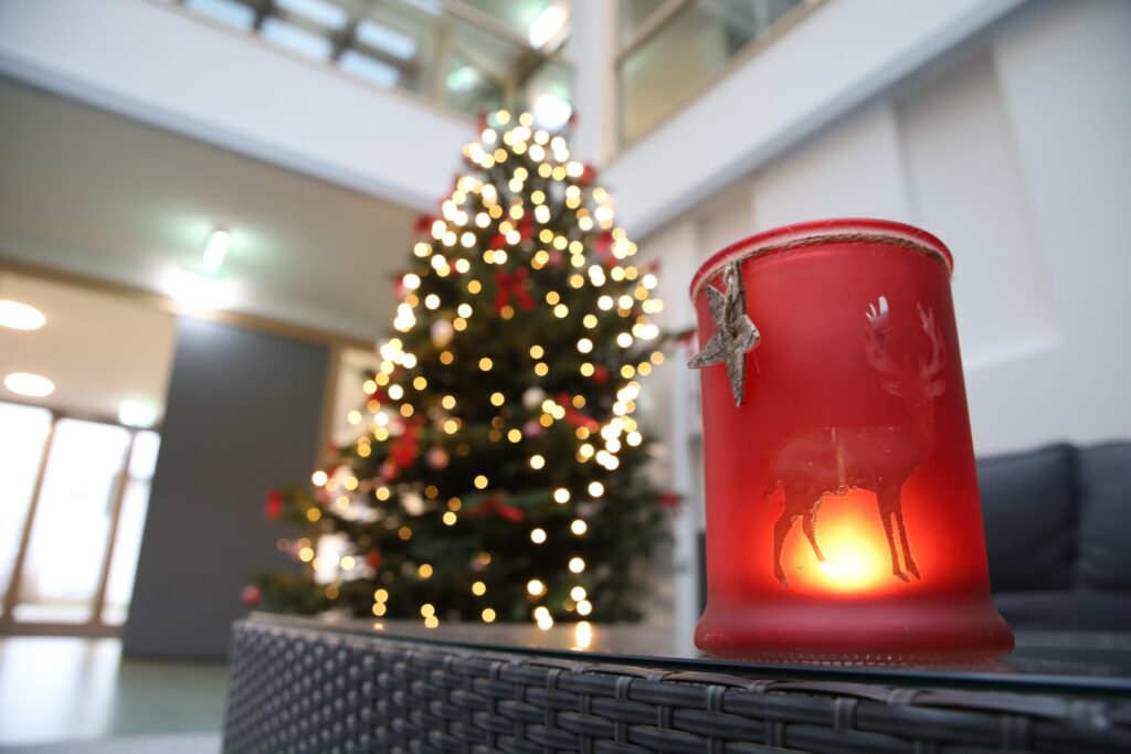 Ein roter Kerzenständer mit einem Rentiermotiv und einer brennenden Kerze im Inneren steht auf einem Tisch. Im unscharfen Hintergrund ist ein geschmückter Weihnachtsbaum mit leuchtenden Lichtern in einer modernen Inneneinrichtung zu sehen.