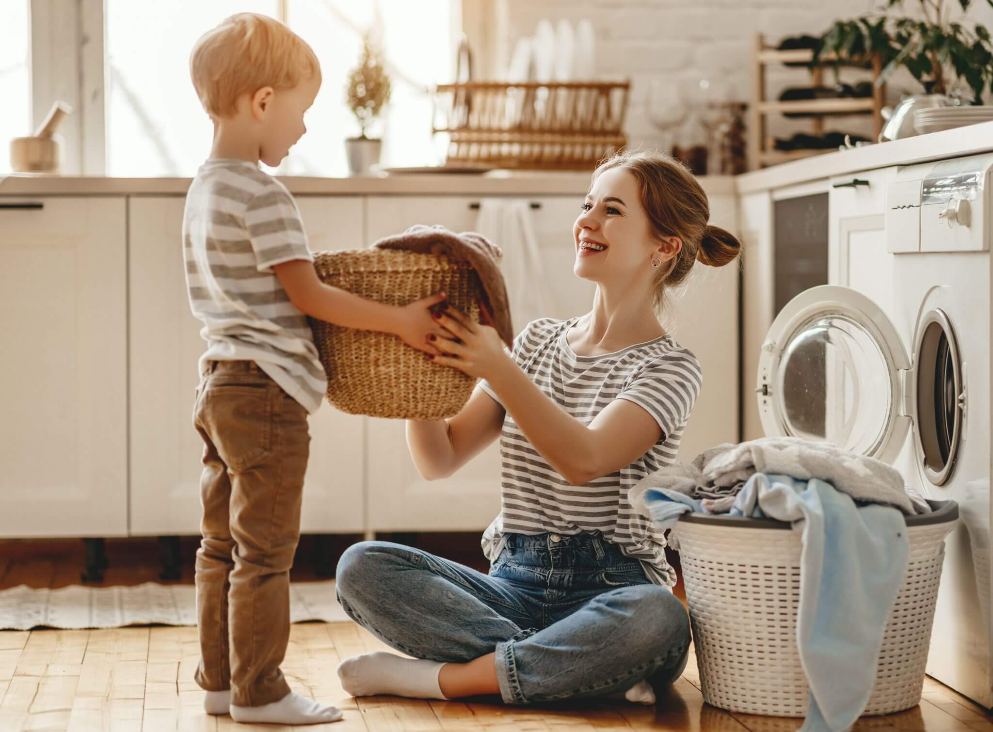 Eine Frau, die auf dem Küchenboden sitzt, lächelt, während sie einem kleinen Jungen hilft, einen Wäschekorb zu halten; eine Waschmaschine und ein Wäschekorb stehen in der Nähe, und beide tragen gestreifte Hemden.