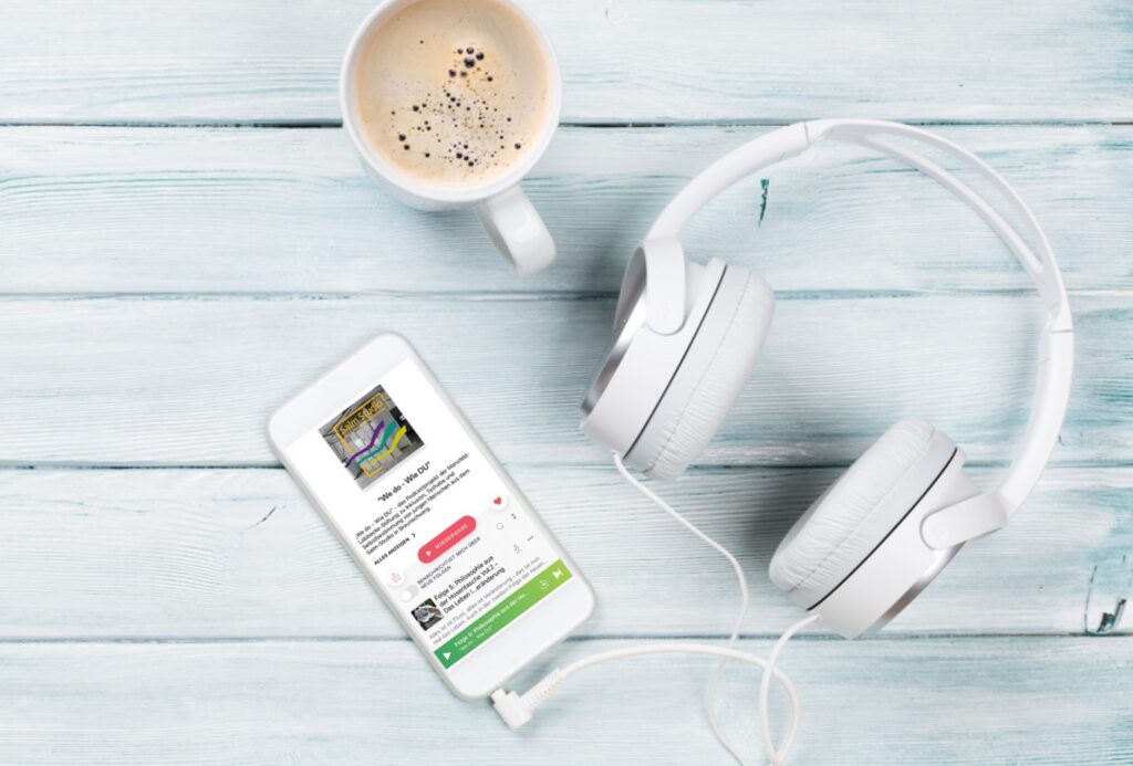Eine Tasse Kaffee, ein weißer Kopfhörer und ein Smartphone, auf dem eine Podcast-Episode angezeigt wird, stehen auf einer hellblauen Holzfläche. Die Kopfhörer sind mit dem Telefon verbunden.