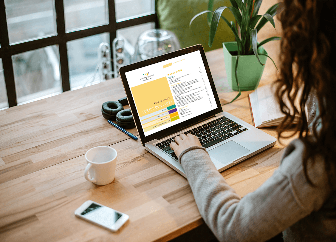 Frau arbeitet am Laptop mit Fortbildungs-Informationen. Homeoffice mit Schreibtisch, Kopfhörer, Tasse und Pflanze.