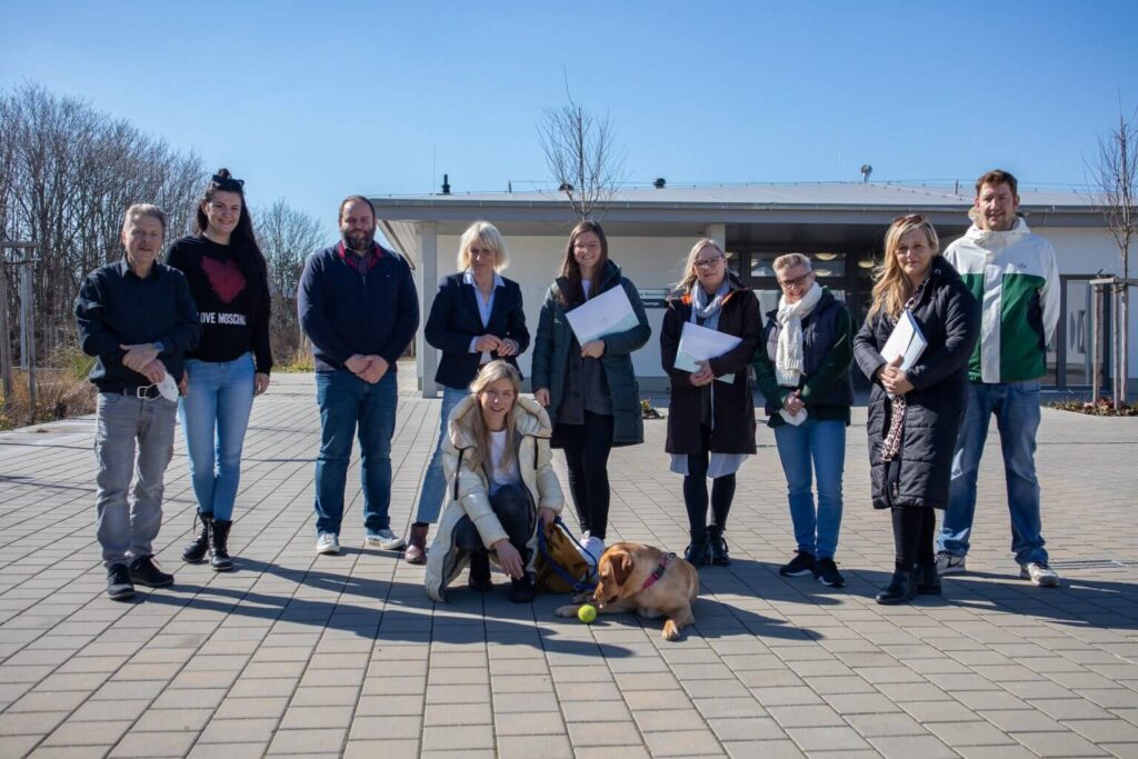 Gruppe von Menschen mit Hund und Tennisball vor Gebäude. Teamfoto, Außentermin, sonniger Tag.