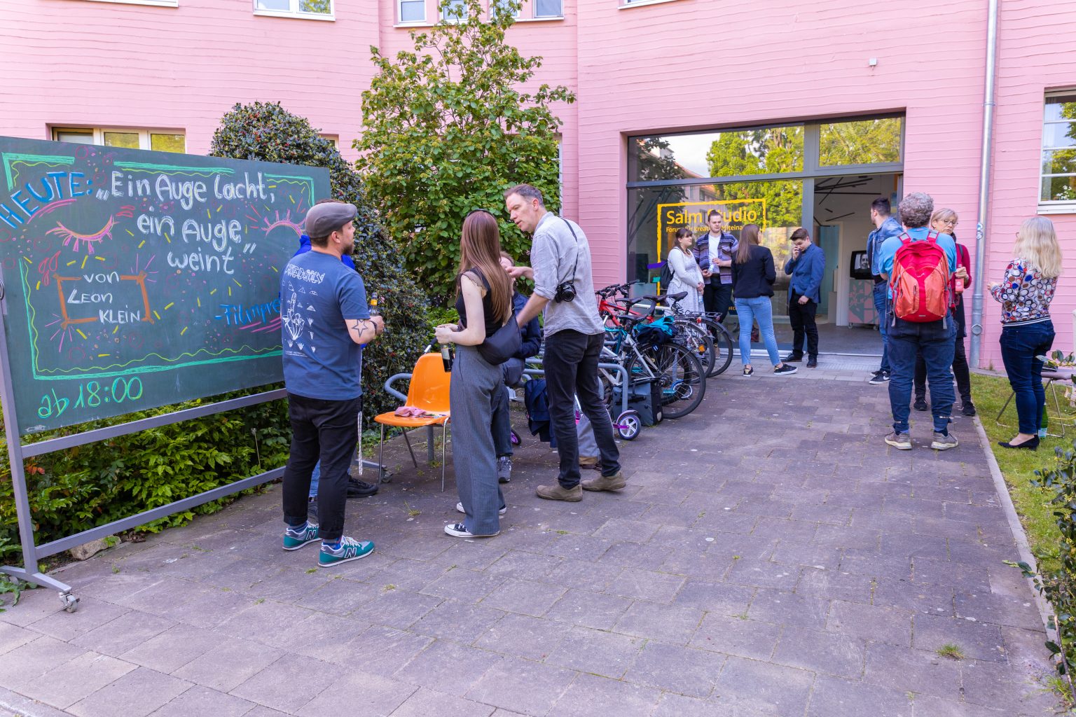 Vernissage "Ein Auge lacht, ein Auge weint" von Leon Klein, Salm Studio. Kunstausstellung mit Besuchern, Fahrrädern und pinkem Gebäude.