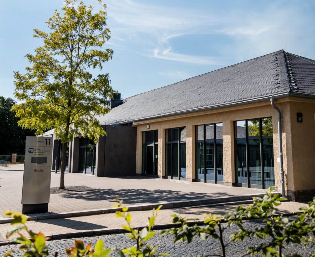 Hörsaalgebäude Universität, Campus mit Baum und Pflastersteinen. Architektur mit großen Fensterfronten und Hinweisschild.