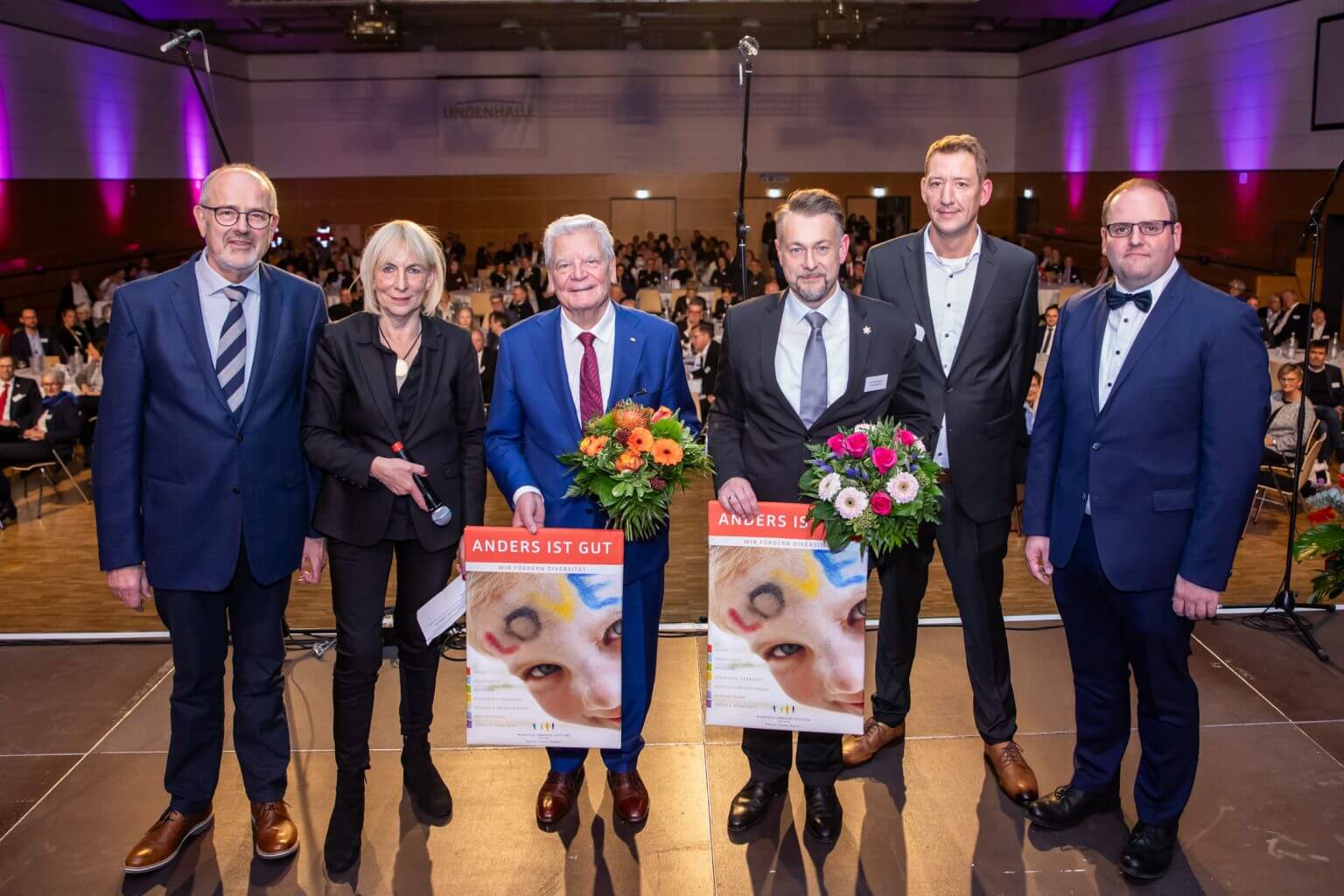 Preisverleihung "Anders ist gut": Gruppe von Personen mit Blumensträußen und Plakaten auf der Bühne, festlicher Anlass.