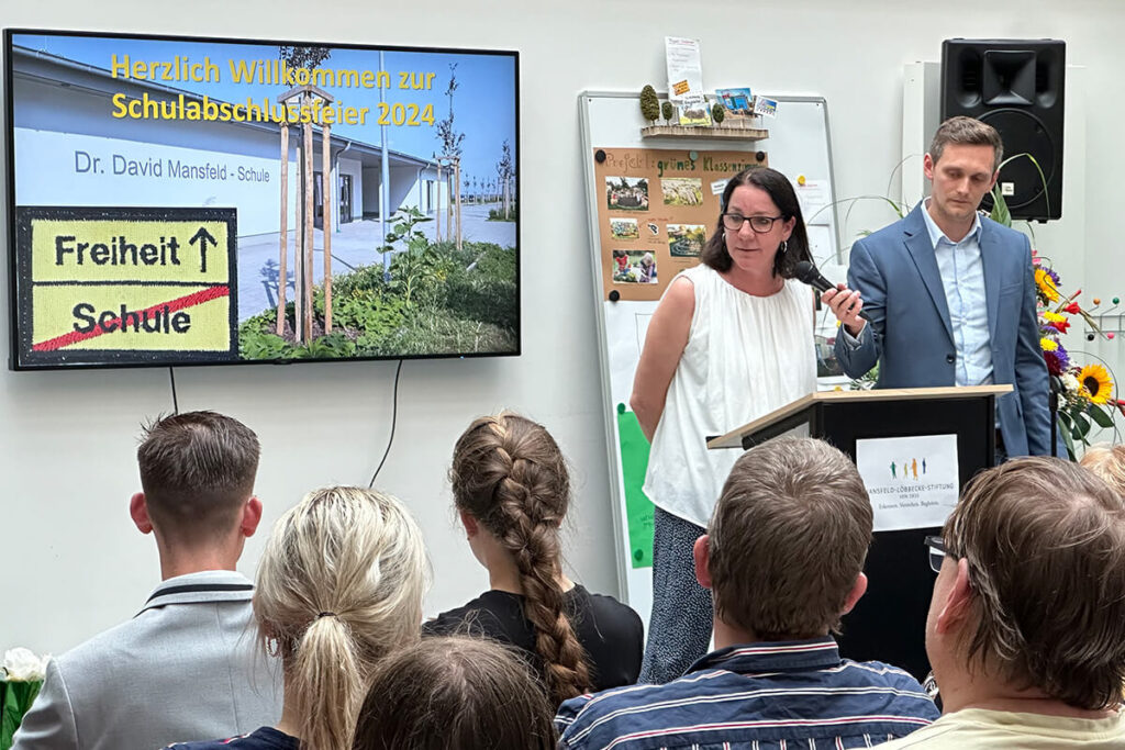 Mann und Frau am Rednerpult, umgeben von einer aufmerksamen Menge.