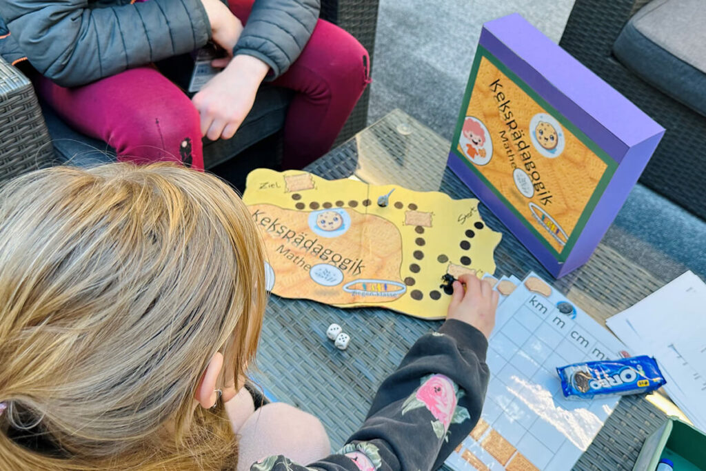 Kind spielt "Kekspädagogik Mathe" Brettspiel mit Würfeln, Oreo-Keksen und Maßeinheiten-Tabelle im Freien. Lernspiel für Kinder.