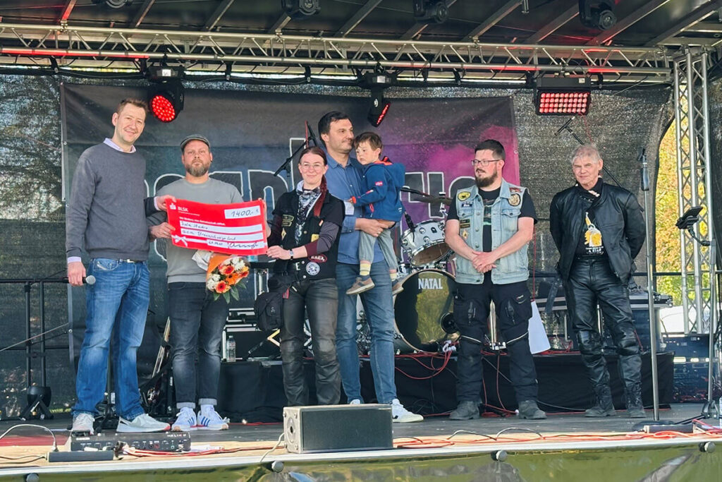 Spendenübergabe auf Bühne: Gruppe von Personen mit Scheck über 1400 Euro, Blumenstrauß, Schlagzeug im Hintergrund. Wohltätigkeitsevent, Musikfestival.