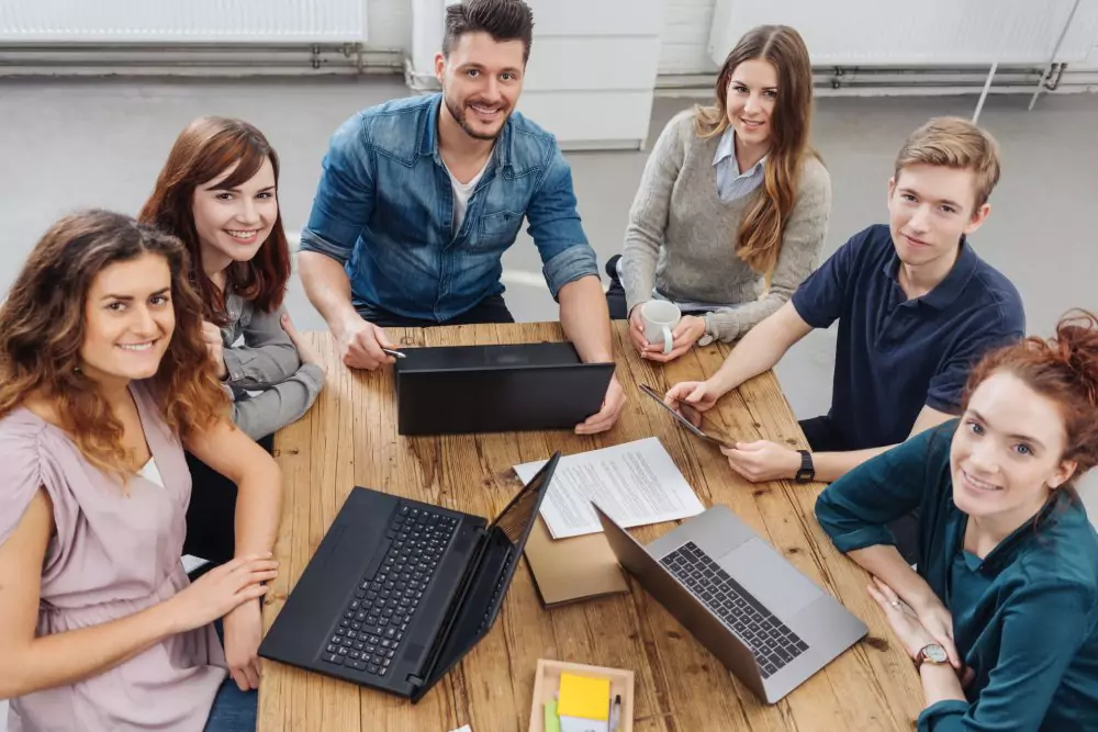 Eine Gruppe von sechs jungen Erwachsenen sitzt mit Laptops und Papieren um einen Holztisch herum und lächelt in die Kamera. Die Atmosphäre ist kooperativ und optimistisch.